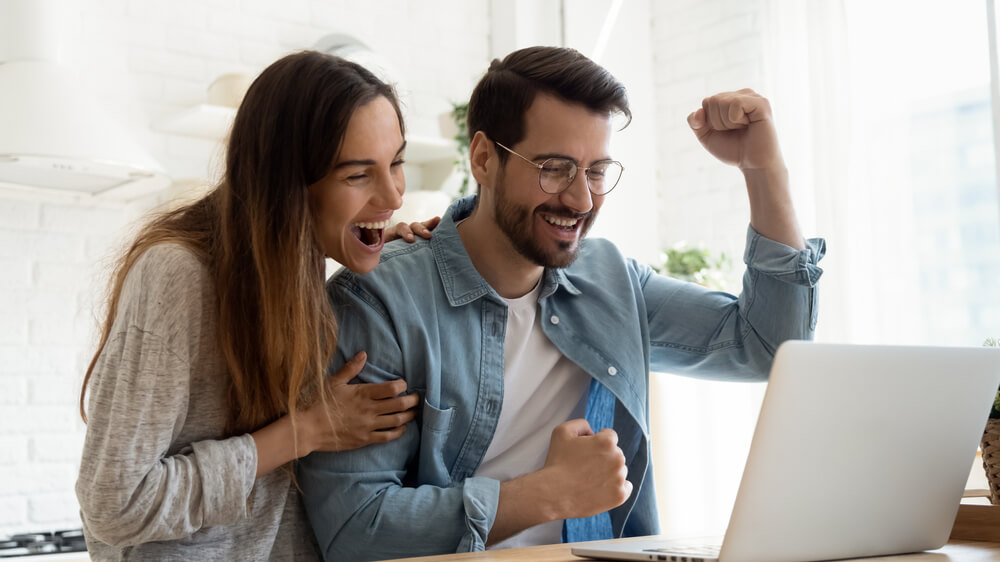 Overjoyed millennial couple sit at desk look at laptop screen feel euphoric with online win, excited happy young husband and wife read good news, get unexpected notification on computer, luck concept
