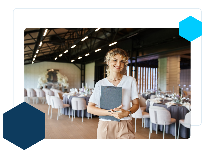 positive decorator with clipboard looking at camera near tables with festive setting in banquet hall