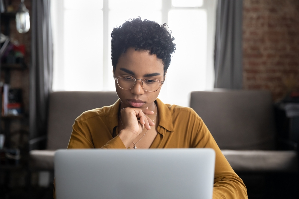 African woman in glasses sit at desk staring at laptop screen