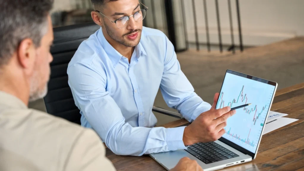 Financial advisor explains stock data consulting to investor. Two busy businessmen conducting financial trading analysis and pointing at the stock market chart on the laptop screen in the office.