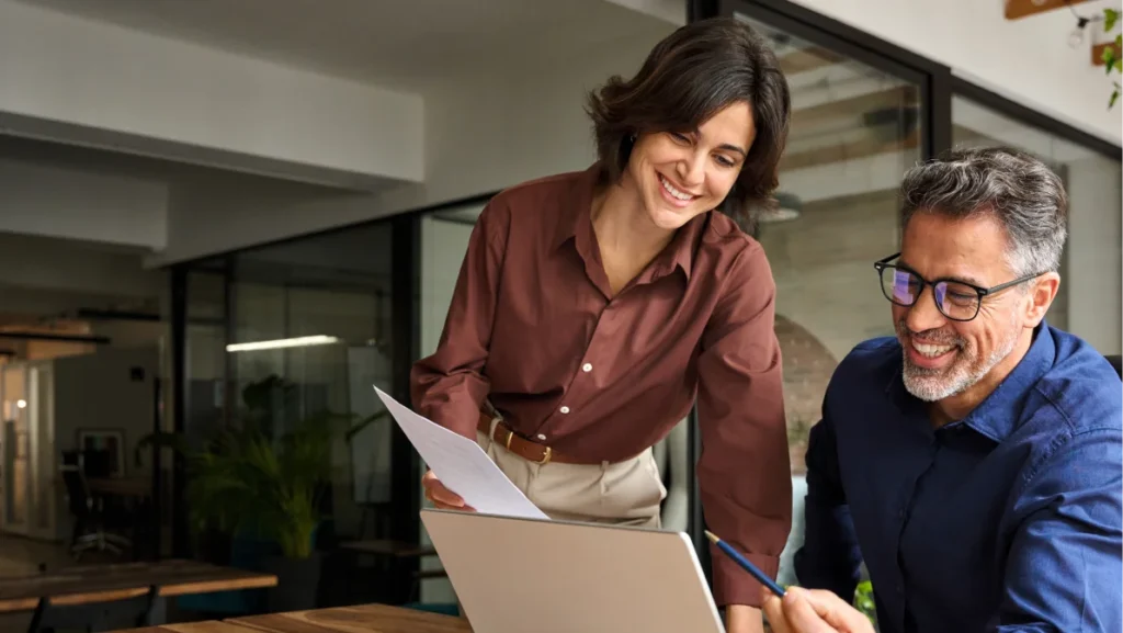 Business team two happy busy executives working together using laptop in office. Young business woman manager showing presentation to mature man investor, consulting client partner at work meeting.
