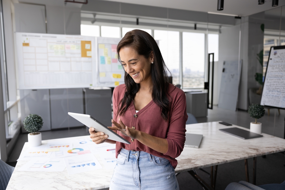 Happy young woman project coordinator type on pad with smile in modern designed boardroom