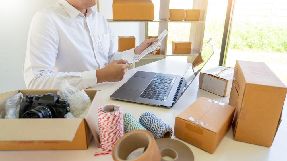 A man packing items in a box, with a laptop and a paper in his hand