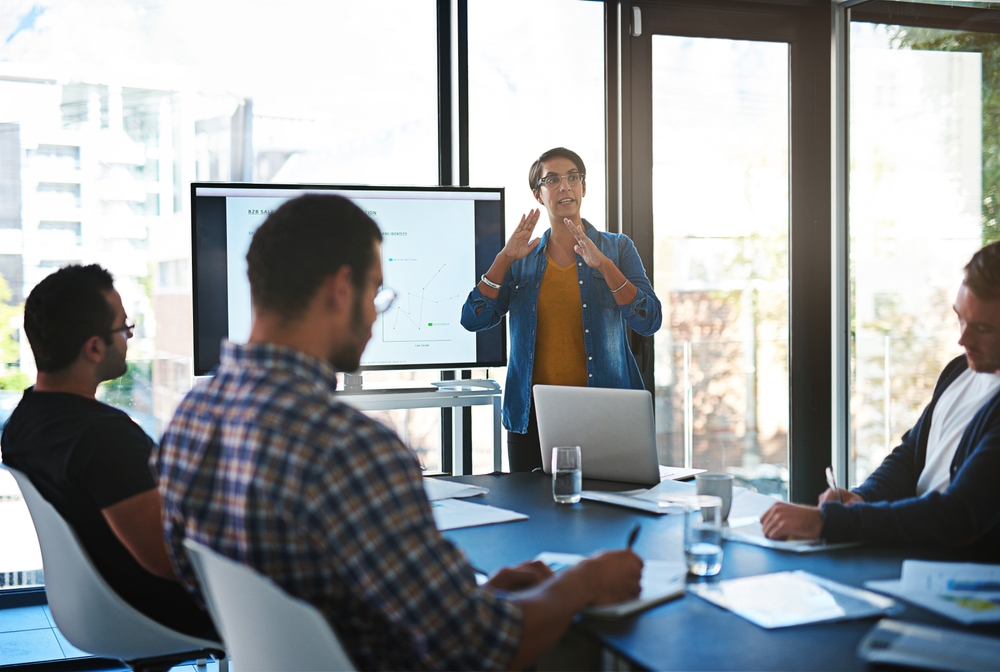 Business woman reporting in front of three men with a screen on her back