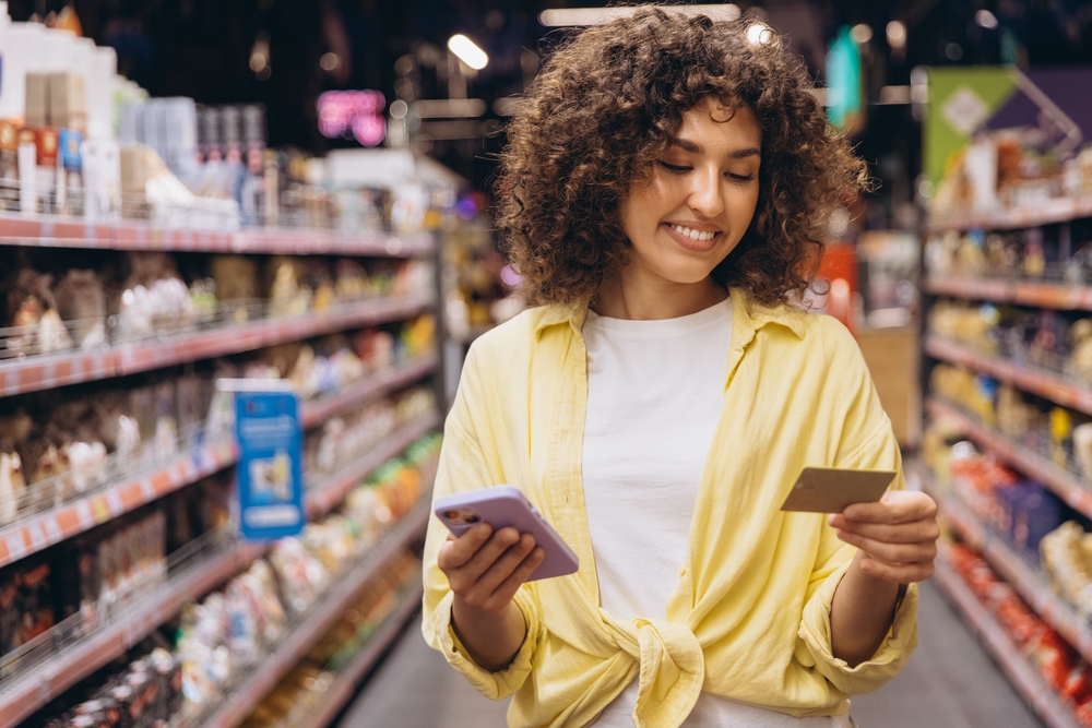 Smiling curly haired woman using smartphone and loyalty card while shopping groceries in a supermarket