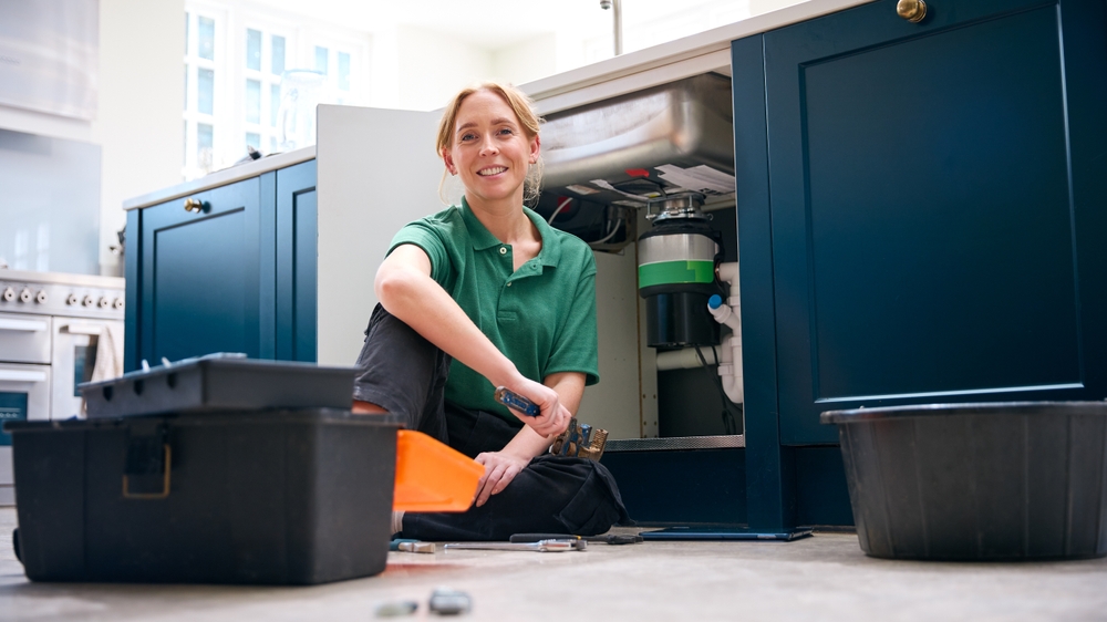 Portrait Of Female Plumber With Digital Tablet Fixing Waste Disposal Unit In Domestic Kitchen Sink