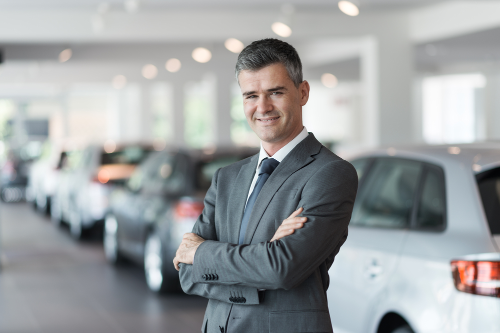 Smiling car salesman at the showroom, standing with arms crossed with luxury cars on the background