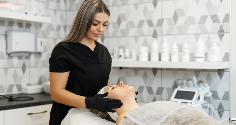 A beautician wearing black gloves performs a facial treatment on a woman lying on a spa bed in a clean, modern clinic with skincare products in the background