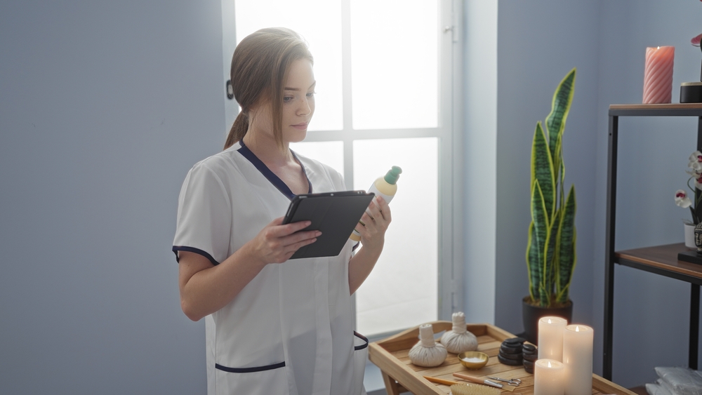 Woman holding tablet and reading product label in spa room with candles and wellness accessories