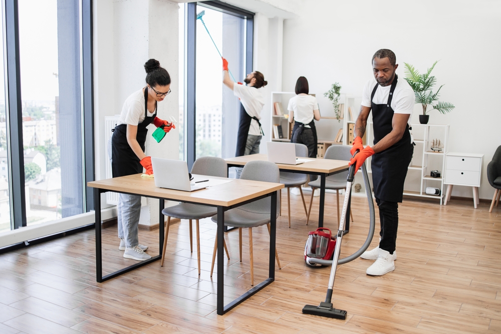 Team of young multicultural cleaners vacuums floor, wipes tables and gadgets, shelves