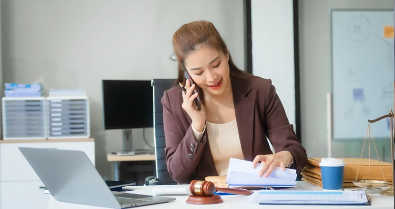 Attractive Asian businesswoman in formal business attire is working at her desk with a laptop and mobile phone, focusing on corporate law, contracts, compliance, and law firm marketing strategies.