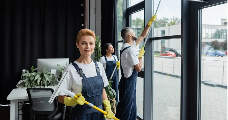 smiling woman in rubber gloves looking at camera near multiethnic colleagues cleaning large office windows