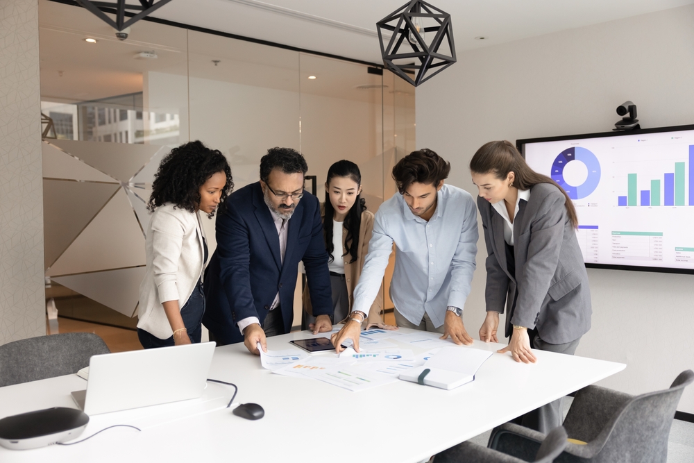 Group of five professionals engaged in collaborative discussion around table in modern office, examining printed charts and data