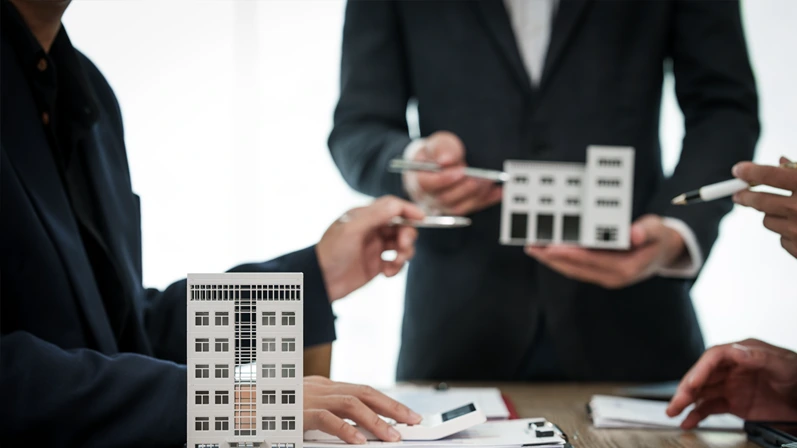 A professional businessman in a suit conducts a meeting at a desk while showcasing a model of a condominium or apartment building, discussing real estate development plans.