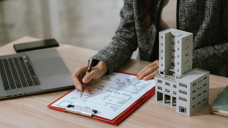 Businesswoman holding a small building model at a desk with a laptop and documents, representing real estate investment, property management, and financial decision-making.
