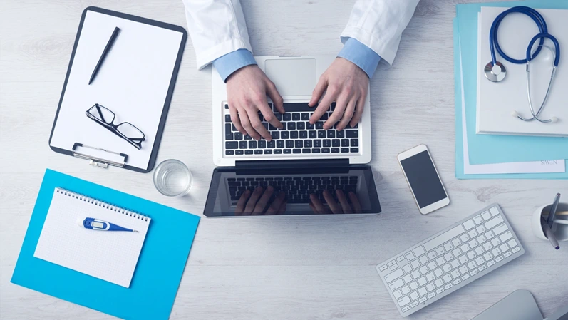 This high quality expertly captured flat lay photograph presents a bird's eye view of a contemporary medical practitioner workspace situated on a pale ivory wood grain surface. The central focus features the skilled hands of a healthcare provider wearing a snow colored clinical coat and a light azure button down shirt as they type efficiently on a metallic laptop keyboard. The desk is meticulously arranged with various essential healthcare instruments and digital tools that suggest a sophisticated blend of traditional medicine and current telehealth practices. To the right of the computer sits a pearlescent smartphone and a silver wireless input device alongside a navy blue stethoscope resting on a stack of sky blue patient files and alabaster folders. On the left side of the frame a charcoal black clipboard holds a neutral document with a dark ink pen and a pair of chic reading glasses placed neatly on top. Below the clipboard is a vibrant cerulean folder supporting a spiral bound notebook with a digital thermometer resting on the clean page. A clear glass of water and a small container of writing pencils are also visible indicating a long and focused workday in a hospital or private clinic setting. The soft diffused lighting throughout the scene creates a sterile yet inviting atmosphere that is perfect for health centered blogs medical technology websites and marketing materials for insurance companies or pharmaceutical brands. This visual effectively communicates themes of precision care organization and the digital transformation of the wellness industry. The thoughtful arrangement provides a balanced composition and refined aesthetic suitable for website hero banners and social media content focused on clinical management. It captures the essence of a dedicated physician managing patient data or conducting remote consultations in a contemporary environment while maintaining a bright and airy visual style.