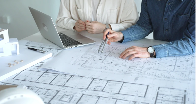 Architects engaging in a professional meeting, reviewing detailed blueprints on a table, utilizing a laptop, and collaborating on building design and construction planning