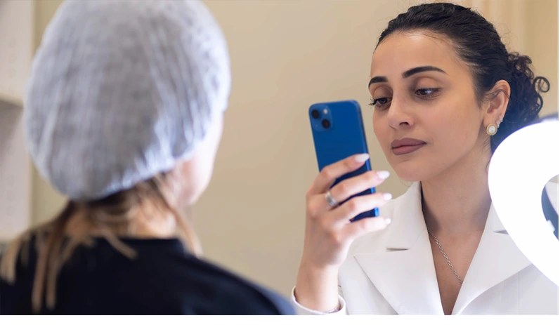 A cosmetologist photographs the results of a skincare treatment using her smartphone under professional lighting. Sharing real-time results on social media reflects modern marketing in the beauty