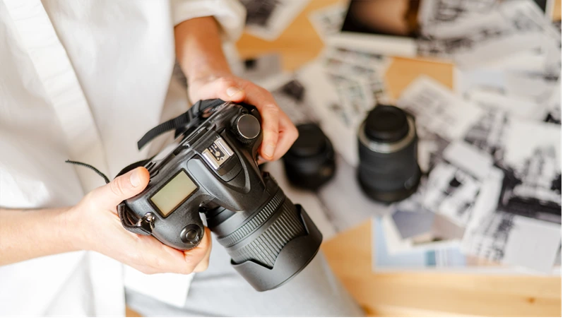 Photographer holding professional digital camera in home office setting, reviewing printed photographs spread out on desk, showcasing photography workflow and creative process