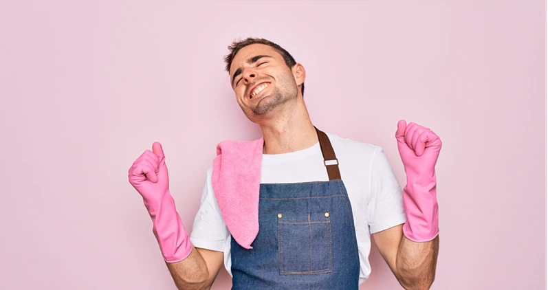 Young cleaner man with blue eyes cleaning wearing apron and gloves over pink background very happy and excited doing winner gesture with arms raised, smiling and screaming for success. Celebration