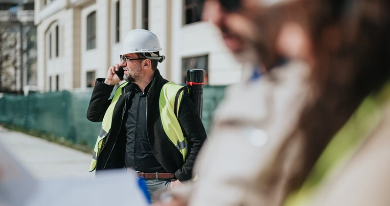 Construction site manager wearing a hard hat and high-visibility vest speaks on a mobile phone. Colleagues in the foreground review plans and documents while the manager oversees the site.