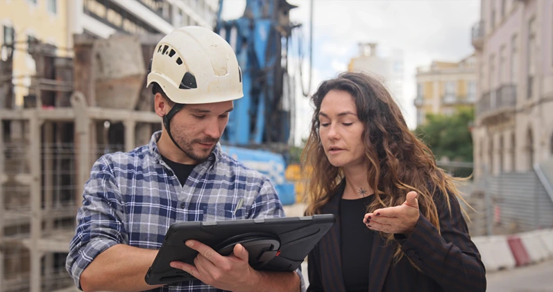 Engineer constructor discuss build plan with investor woman. Builder guy show construction site project tablet pad. Worker tell blueprint client. Man work city street. Customer look new house scheme.