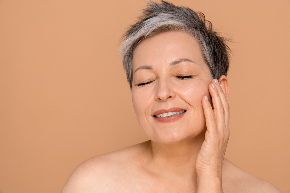 Gorgeous senior older woman with closed eyes touching her perfect skin.