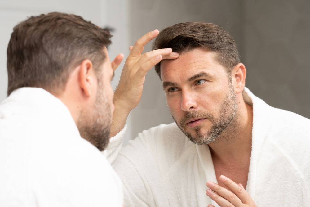 Middle-aged man carefully inspects wrinkles and skin texture in bathroom mirror.