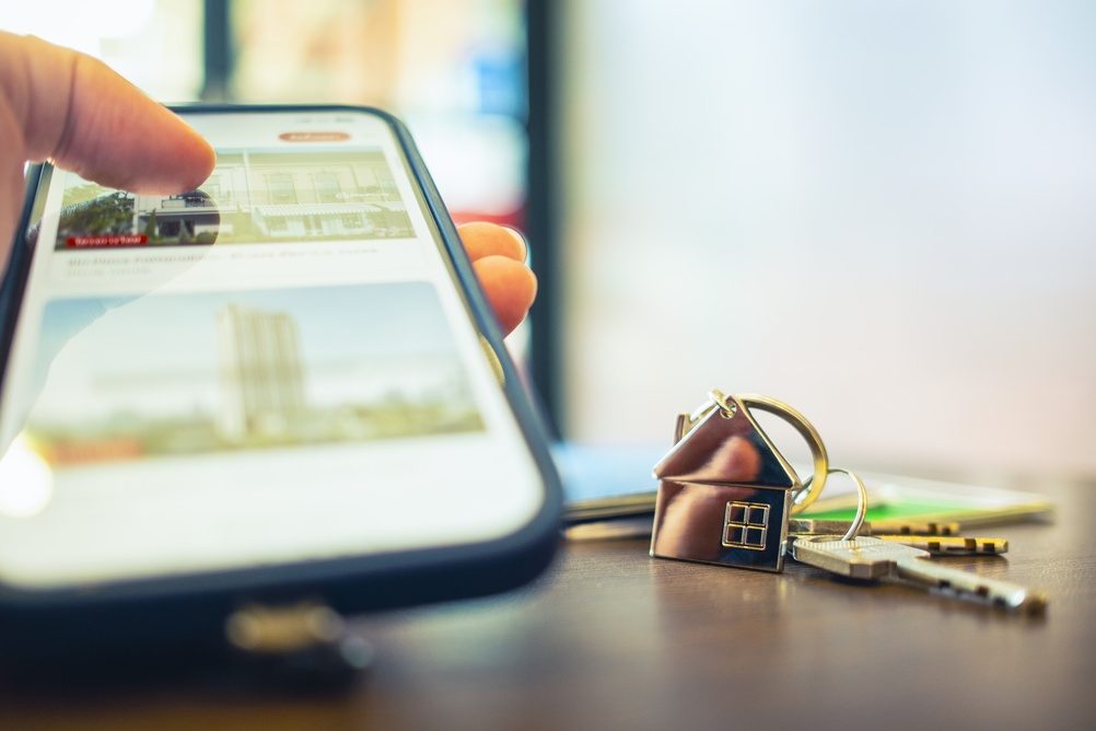 Close up of hand using smartphone searching for real estate house on screen with key on table. 