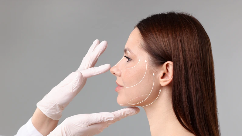 Woman preparing for plastic surgery, grey background. Doctor examining her face with markings, closeup
