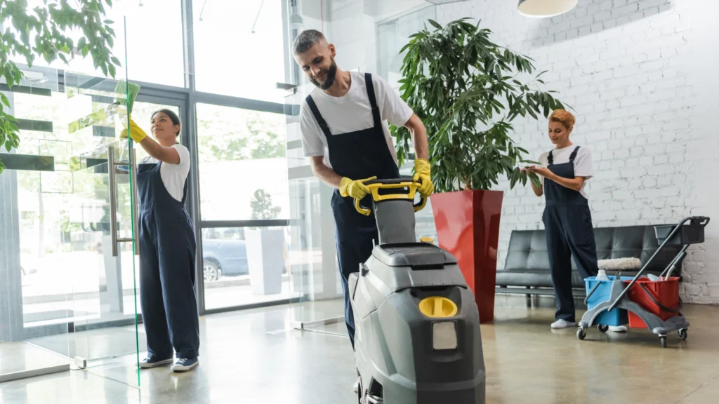 man in workwear with floor scrubber machine near interracial women cleaning office lobby