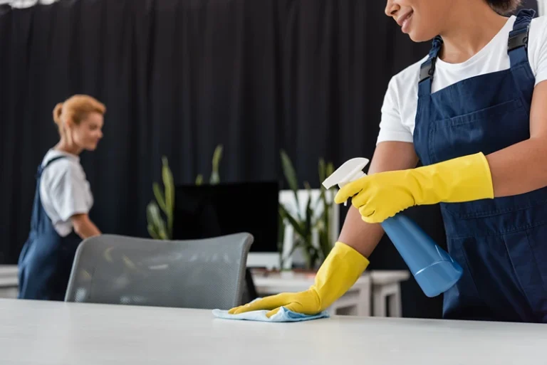 smiling bi-racial woman cleaning office desk with detergent and rag near blurred colleague