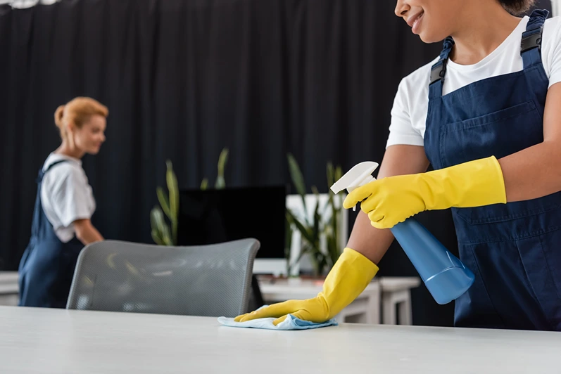 smiling bi-racial woman cleaning office desk with detergent and rag near blurred colleague