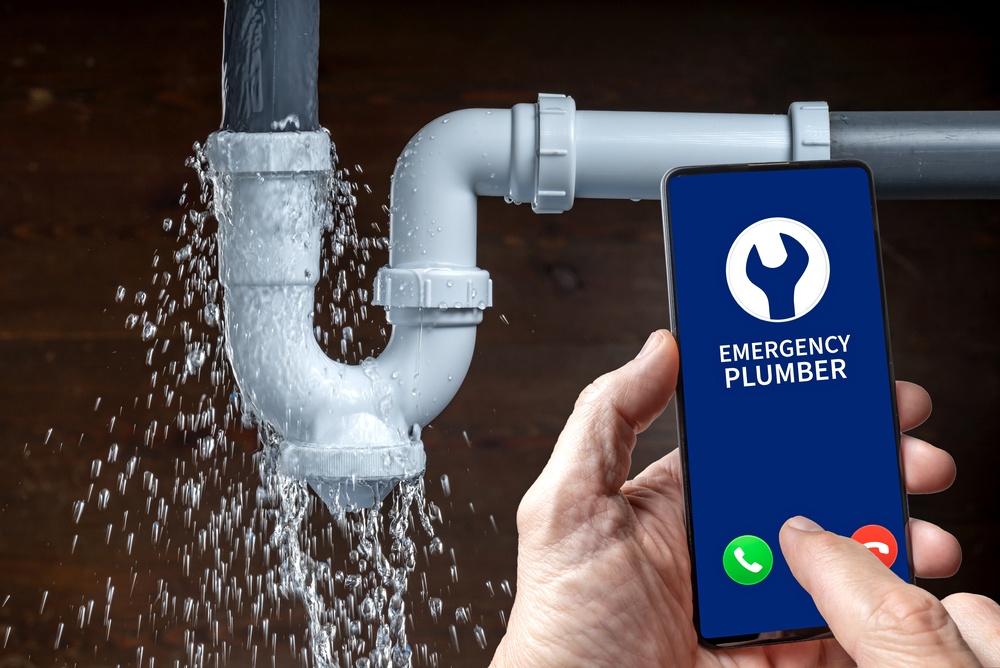 Leaking sink pipe and emergency plumber call on smartphone for home repair service.